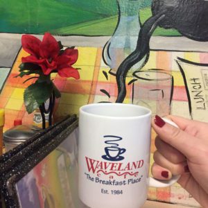 White coffee cup reading "Waveland Cafe" in front of coffee pouring mural at the Waveland Cafe in Des Moines, Iowa