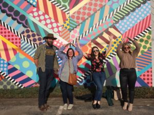 Four people standing in front of a mural spelling Iowa with their arms on a tour with the Iowa Tour Company in Des Moines, Iowa