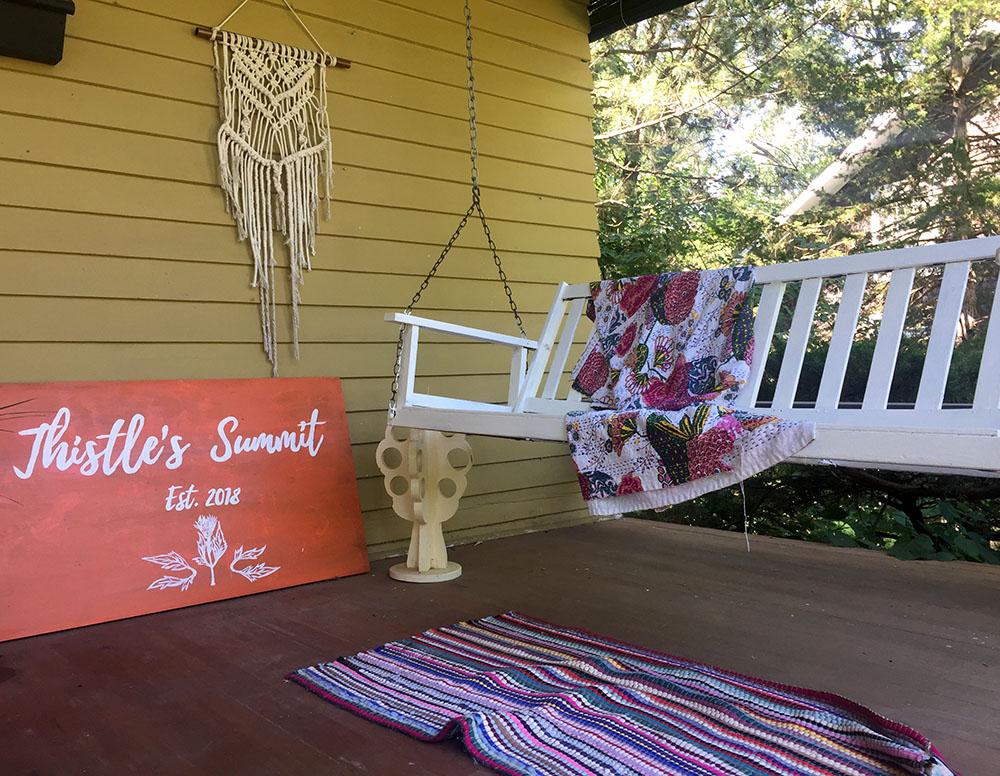 White front porch swing with colorful blanket and orange sign reading "Thistle's Summit" at Thistle's Summit in Mount Vernon, Iowa