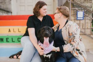 Two people with a black dog in front of rainbow painted stairs in Mount Vernon, Iowa