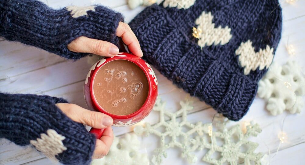 Hands wearing navy gloves holding a red mug of hot cocoa against a white snowflake background | Olio in Iowa's 2020 Holiday Gift Guide
