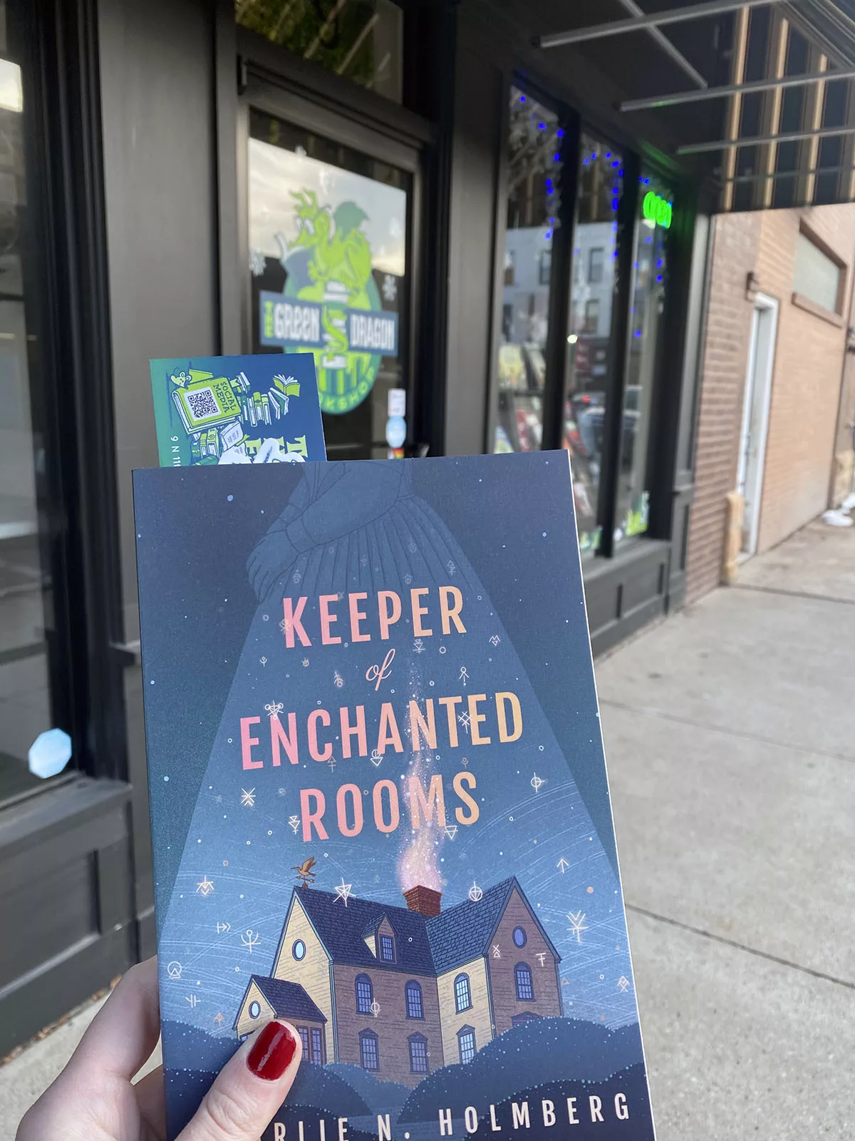 Hand holding book titled Keeper of Enchanted Rooms in front of Green Dragon Bookshop in Fort Dodge, Iowa