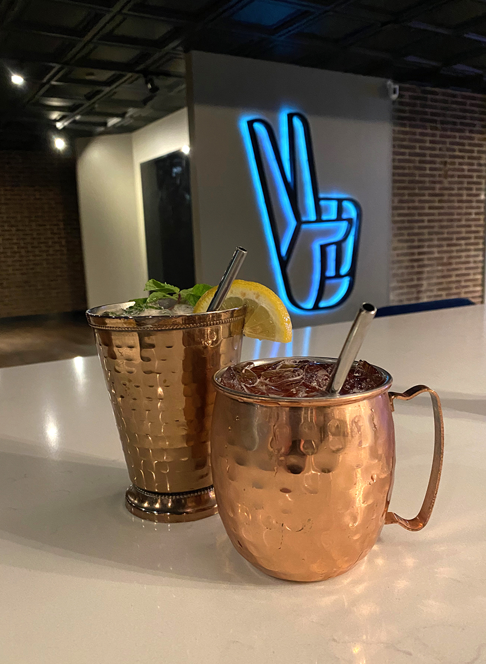 Two drinks in copper mugs on a bar with a blue backlit peace sign sculpture in the background