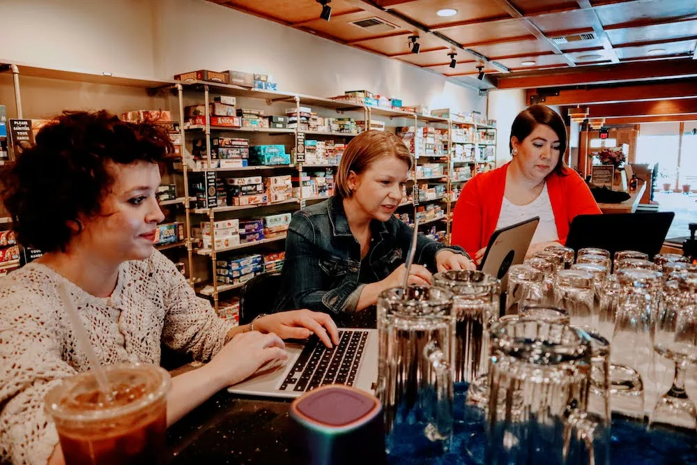 Three women sitting at a bar using laptops with board games in the background