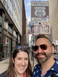 Two people standing in front of Historic Begin Route 66 sign in downtown Chicago on E. Adams Street and Michigan Avenue