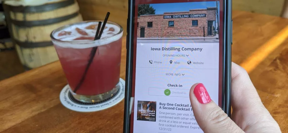 Hand holding phone using the Iowa Distillery Pass with a cocktail in the background at Iowa Distilling Company in Cumming, Iowa