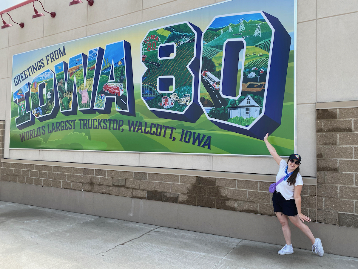 Woman standing in front of mural that says "Greetings from Iowa 80" at the World's Largest Truckstop in Walcott, Iowa