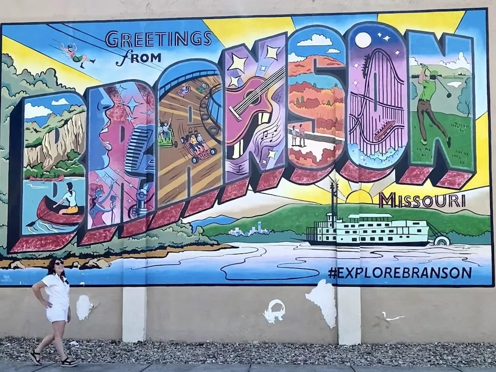 Woman walking in front of colorful mural that says "Greetings from Branson" in Branson, Missouri
